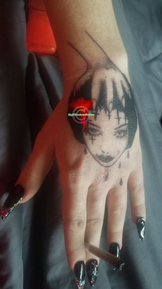 High Priestess Kiefsten's first tattoo! The tattoo is on her hand, and is a goth woman's head decapitated & dripping blood, being held in a clawed hand. All bold black with some white highlights for the eyes & lips. 

High Priestess Kiefsten's hand has painted, stoned nails & is holding a joint.