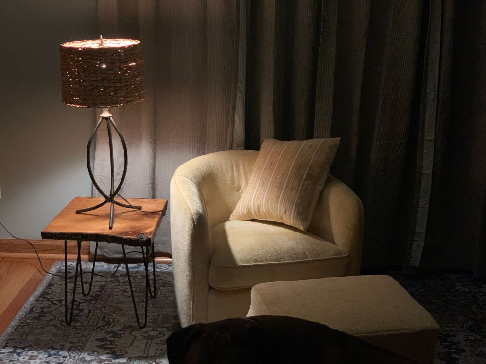Hey raw edge table with hairpin legs. A metal lamp base with a grass shade sitting behind a light yellow, swivel chair and stool with a coordinating pillow. 