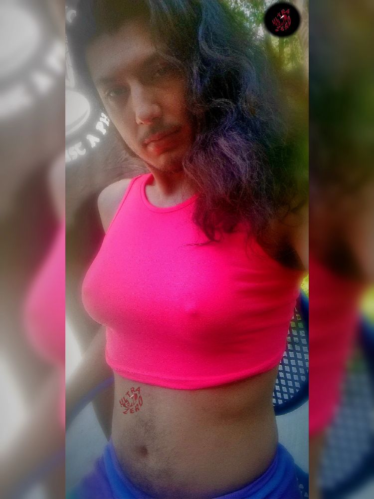 Selfie of UltraEvilZero with his hair down no glasses, wearing a pink sleeveless crop top with fake Titties inside them with colorful purple pj's on as he sits outside 