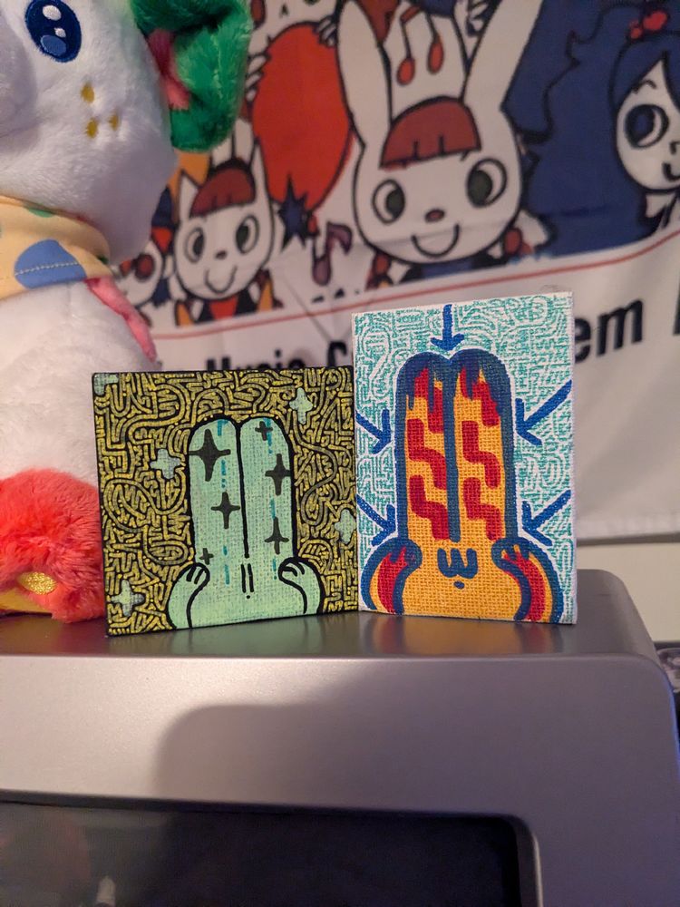 a picture showing two small canvases sitting on a CRT tv, with a plushie next to it and a popn music flag on the wall behind. they show drawn, stylized rabbits in a mosaic like pattern. fine art!
