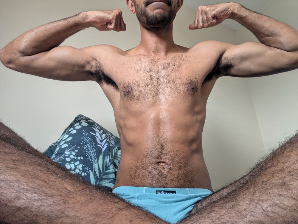 Man sat on a bed in his underwear, flexing both arms.