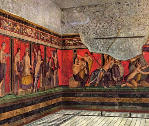 A corner of a room at the Villa of the Mysteries at Pompeii from the 1st century BCE. There's a wall painting with lots of people doing inscrutable things on a bright red background. It's an initiation into the Dionysian mysteries, probably.