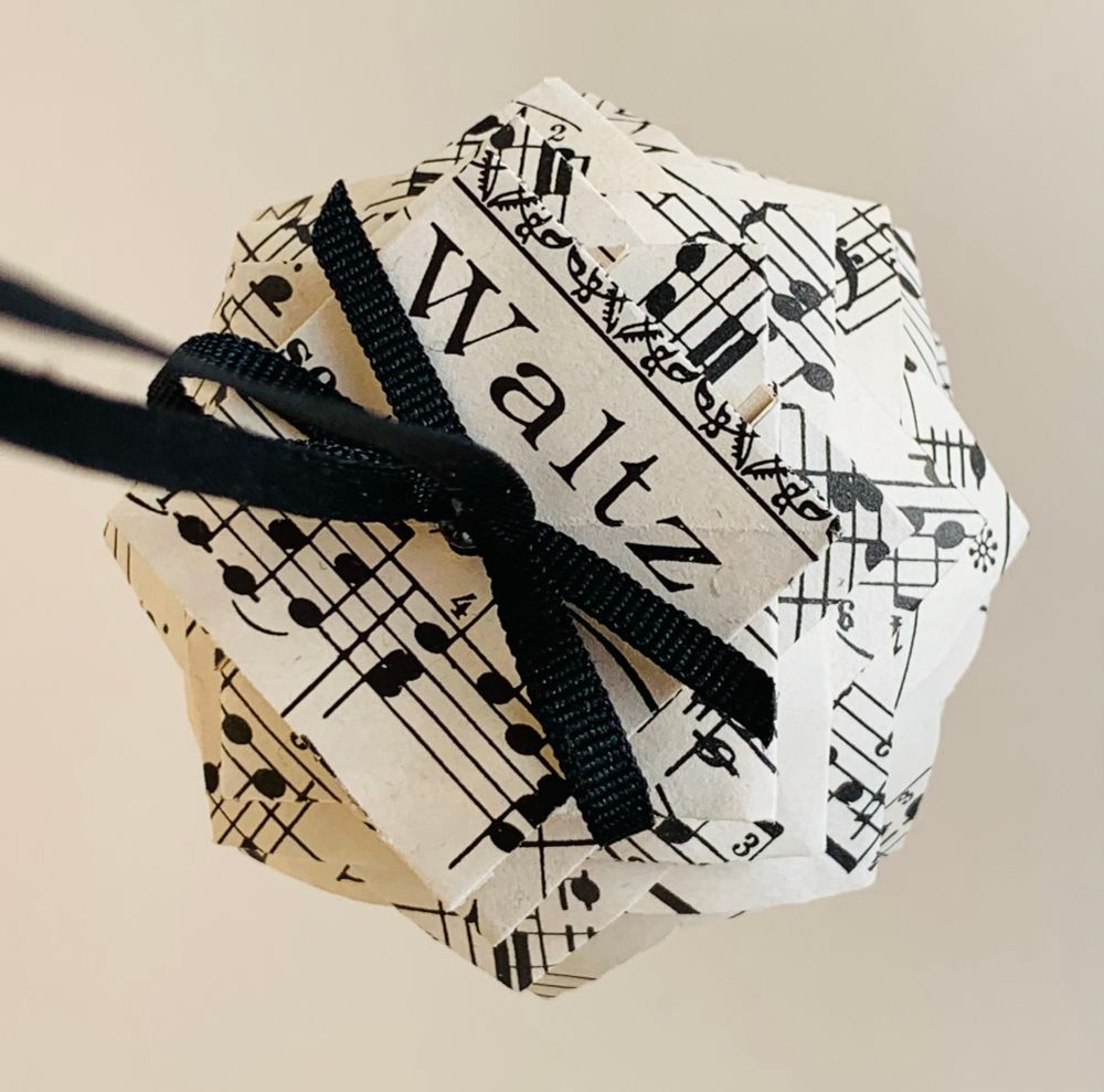 The top of a pinecone ornament made from vintage piano sheet music. The title “Waltz” is in large letters just above the black ribbon bow pinned to the top of the ornament.