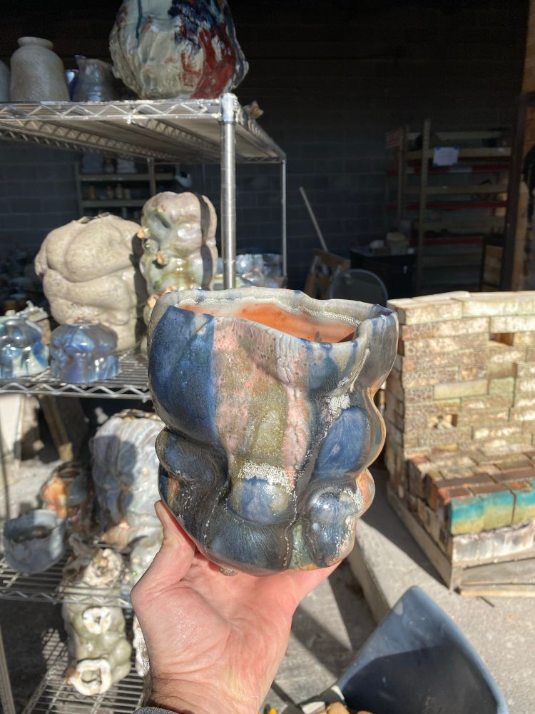 A wood-fired ceramic vessel with glazes and undulating contours, held in hand.