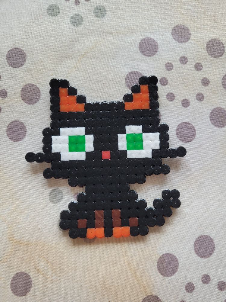 Posé sur la planche à repasser un chat noir aux yeux vert fait en perles à repasser. Son petit nez est rouge. 
