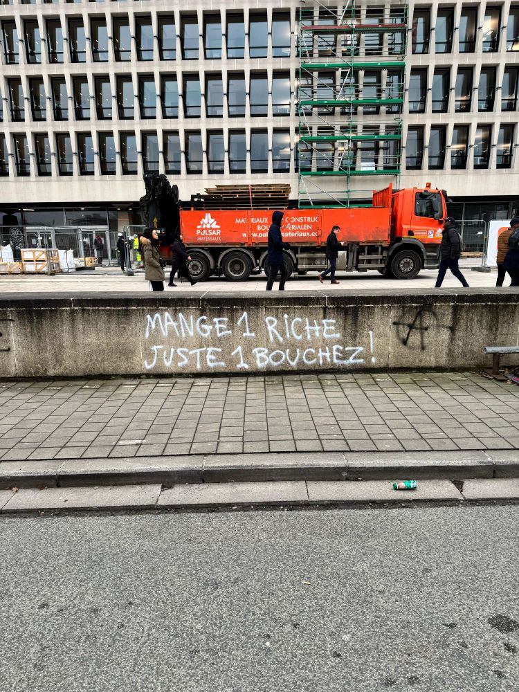 Photo d'un tag blanc sur un muret qui dit "mange 1 riche, juste 1 bouchez". Bouchez est écrit avec un Z, comme le nom de famille de Gloub, Georges-Louis Bouchez.
À côté, un tag en noir d'un A entouré, symbole de l'anarchie.
Derrière le muret, des gens marchent devant un camion orange. 