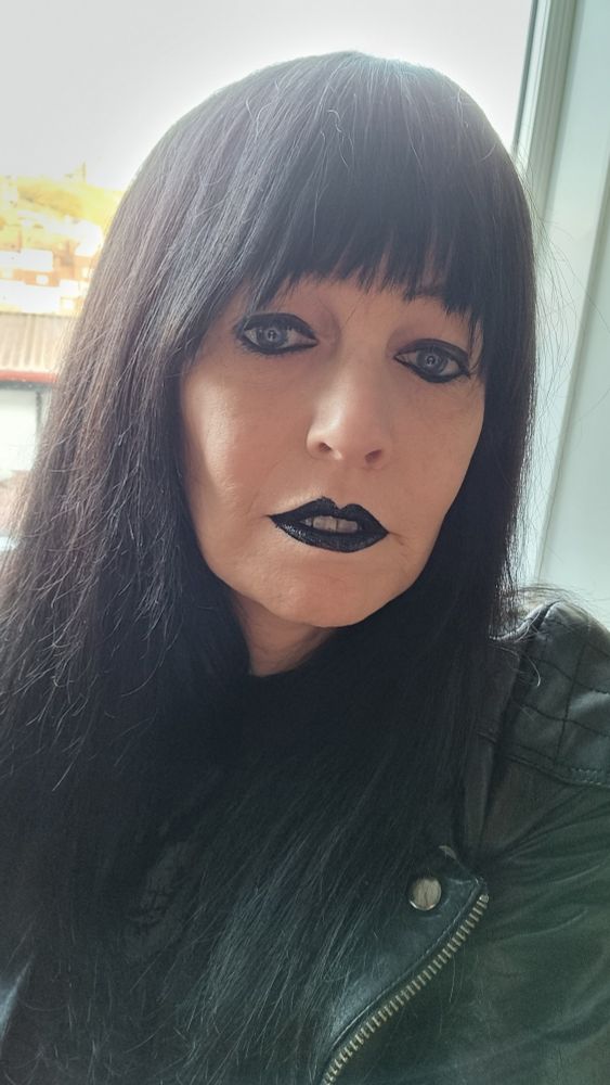 Me,  with black hair, fairly heavy black eyeliner and black lipstick. I'm wearing a black dress and black leather jacket. In the background is a hillside bathed in sunlight, but it's actually a view across Whitby harbour up to the Abbey 