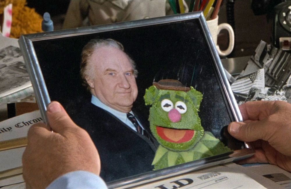 a framed photograph of a man and a muppet is held in human hands. The muppet is a bear with a hat (like Fozzie), but it's green with ping-pong eyes and a pointy frill around its neck (like Kermit).