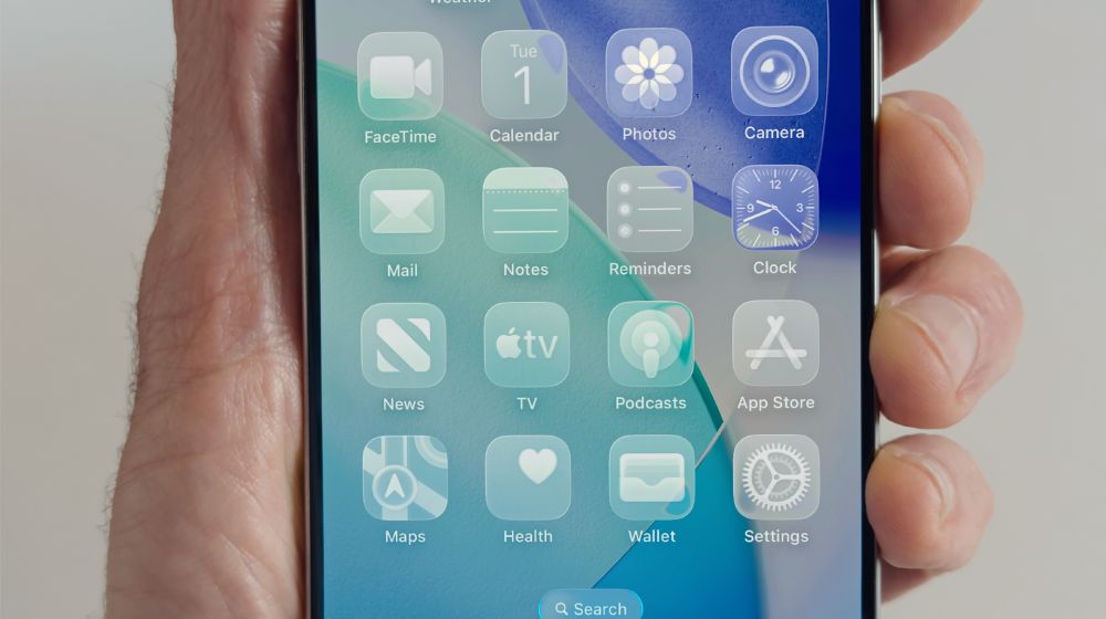 Close-up of a hand holding an iPhone showing the home screen. The icons are displayed in a soft, frosted glass style.