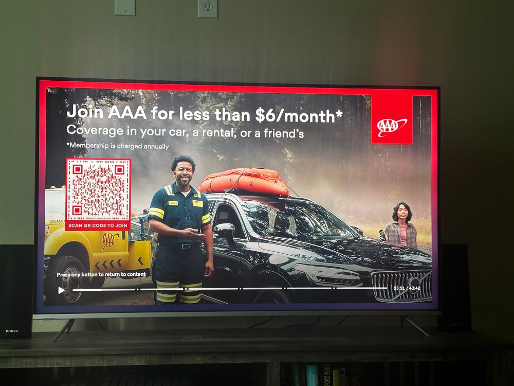 On a TV, the authors photo depicts a pause screen from the Peacock streaming service for an Ad from AAA. A red border covers half of it, woods / fog are in the background, a man, named Lucas, is smiling creepily at the camera while their AAA yellow tow truck is in no position to tow the offbrand Volvo that seems fine. A woman in the left of the screen stares dead eye with her flip screen phone from 2005 