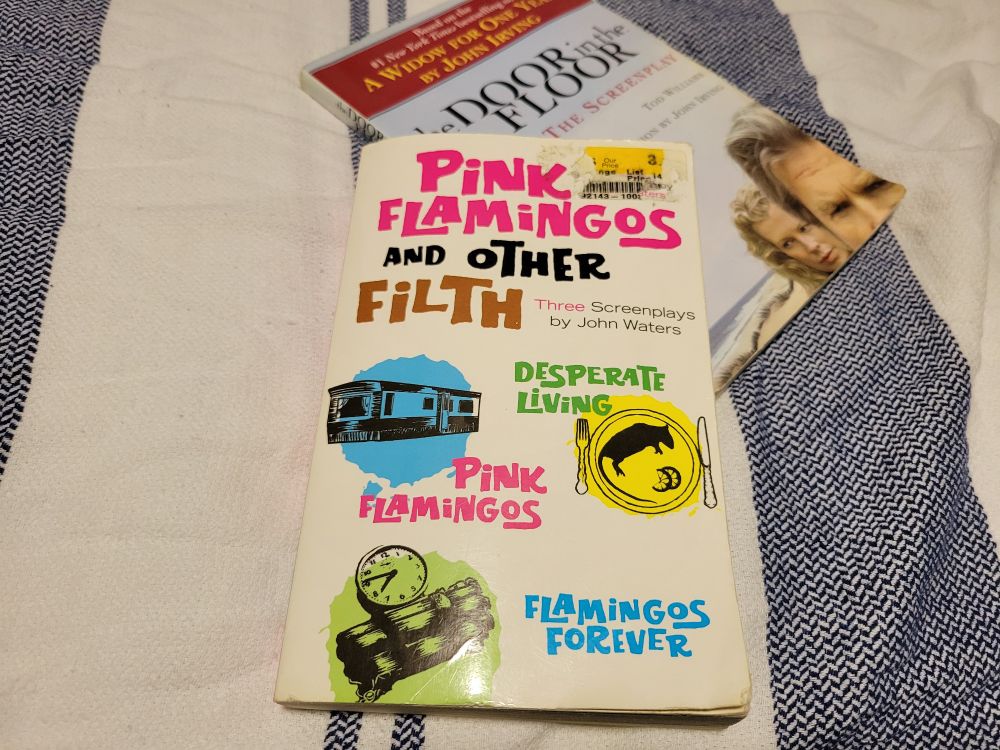 Two paperbacks stacked on a striped towel. The top book is Pink Flamingos and Other Filth: Three Screenplays by John Waters, its cover loud with pop-art circles showing a trailer, a pig, and a clock. Beneath it peeks The Door in the Floor: The Screenplay by Tom Perrotta and Tod Williams, with the faces of two actors framed close together.
