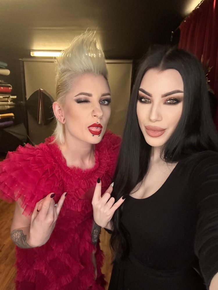 Photoshoot candid/behind the scenes photo of gothic model Ulorin Vex (on the left) and model/photographer Morgana aka Threnody in Velvet (on the right). Ulorin is wearing a red ruffled dress and has blonde hair in a mohawk, Morgana is in all black with long straight black hair. They are both throwing horns. Ulorin is winking and sticking her tongue out