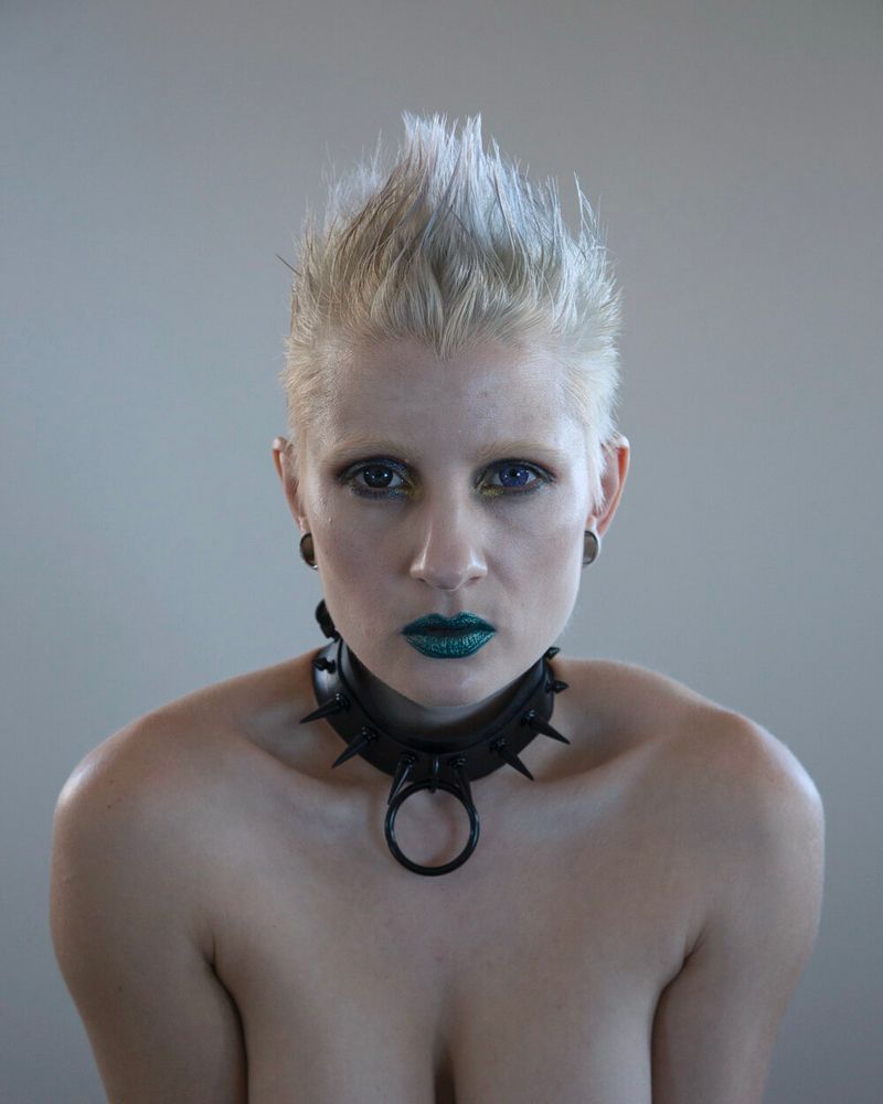 Self portrait head and shoulders photo of artist Ulorin Vex, leaning forward with head face on to camera. She has short spikey platinum hair and is wearing teal lipstick and a black spiked O-ring collar