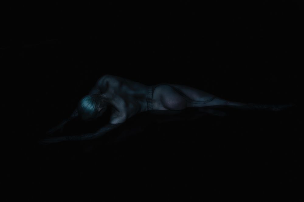 Art nude. Very dimly lit image of a muscular nude female model laying on her side, back to the camera, half submerged in water. It is so dark the water appears black and only the body and face are only partially visible. The model has a large snake tattoo wrapped around the torso