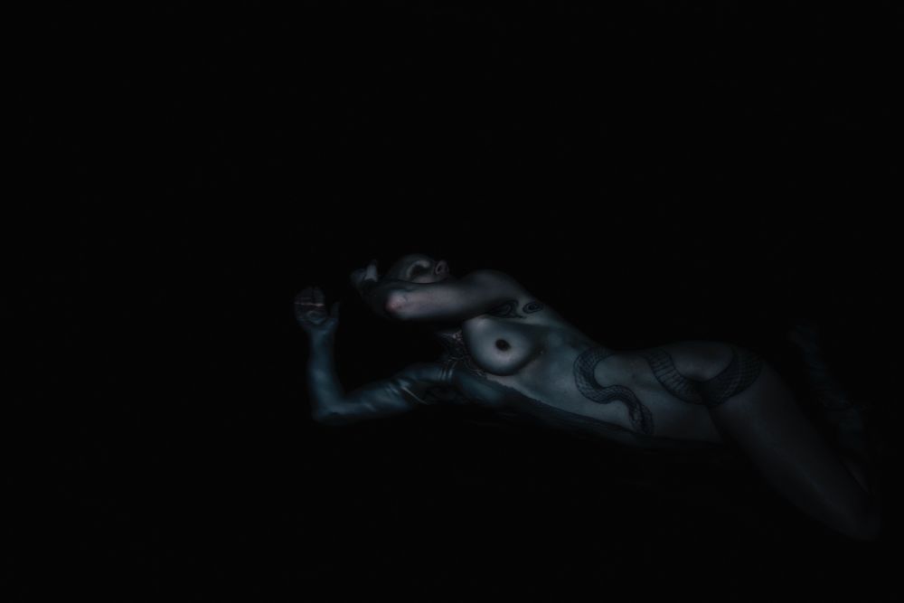 Art nude. Very dimly lit image of a nude female model laying on her side, half submerged in water. It is so dark the water appears black and only the torso and arms are partially visible. The model has a large snake tattoo wrapped around the torso