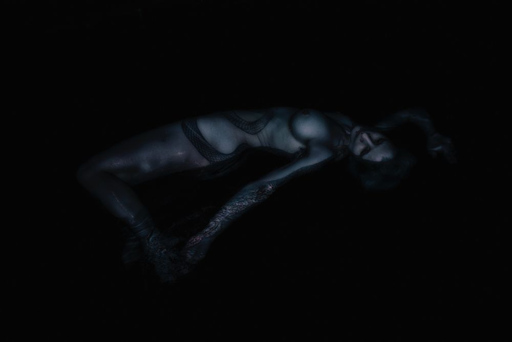 Art nude. Very dimly lit image of a nude female model laying on her back, half submerged in water. It is so dark the water appears black and only the body and face are only partially visible. The model has a large snake tattoo wrapped around the torso