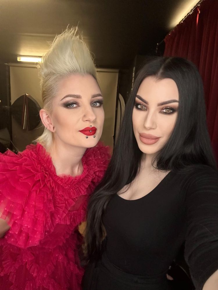 Photoshoot candid/behind the scenes photo of gothic model Ulorin Vex (on the left) and model/photographer Morgana aka Threnody in Velvet (on the right). Ulorin is wearing a red ruffled dress and has blonde hair in a mohawk, Morgana is in all black with long straight black hair