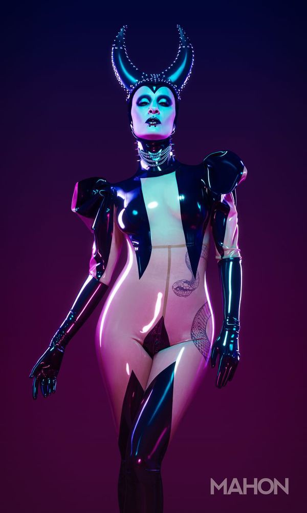 Caucasian model with slim curvy figure wearing a very shiny transparent fully covered latex catsuit. There is black latex appliqué covering forearms, legs up to the thigh, crotch and breasts and it has pointed shoulders. She is also wearing a black latex hood with open face, gothic makeup and a headdress that is reminiscent of Maleficent.

The image is shot against a background with a dark violet gradient that is lighter at the bottom and there are magenta highlights