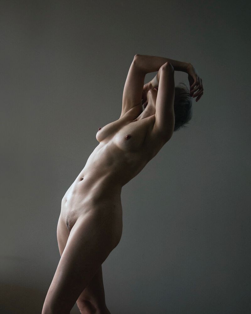 Athletic nude female model standing 2/3 on to camera leaning backwards slightly with arms resting above her head and covering her face. She has short hair which is in shadow. The image is shot in muted desaturated colour with a subtle sepia tone. It is lit with a soft side light from the left with a vignette