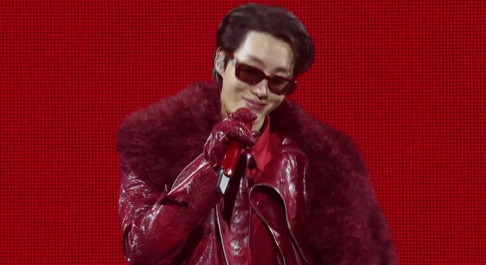 j-hope in a red fur trimmed leather jacked and red leather gloves, holds a red mic and is smiling, wearing sunglasses and standing against a red backdrop