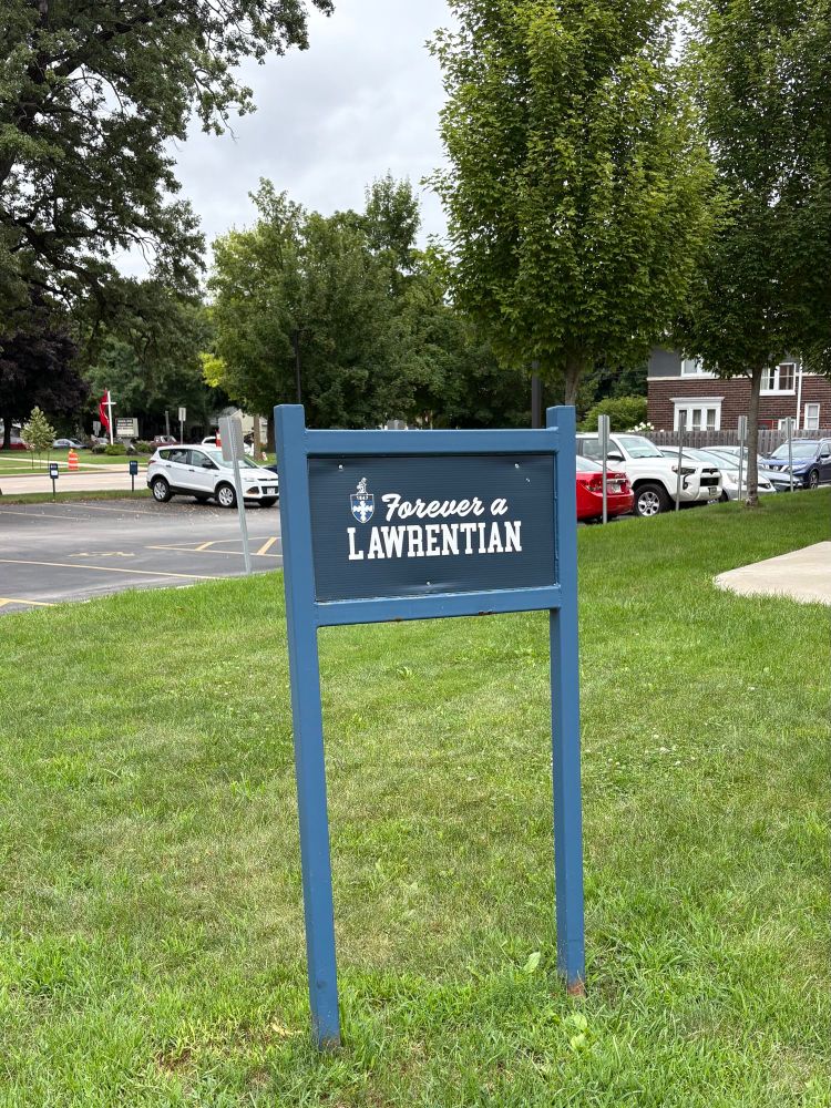 Photo of a sign standing in grass. There is a parking lot in the background. The sign states “Forever a Lawrentian.” 