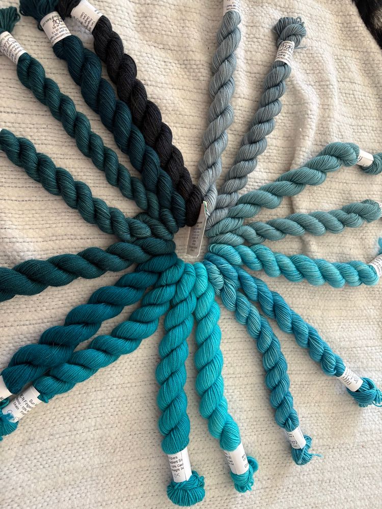 Photo shows 16 mini skeins of yarn in a blue-green gradient arranged in a circle. 