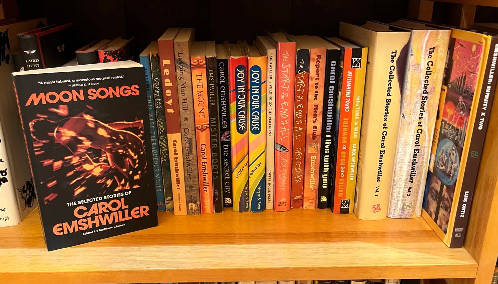 A book called MOON SONGS: The Selected Stories of Carol Emshwiller face out on a shelf of other books by Carol Emshwiller. Moon Songs has a cover with a black background and orange lettering and in the middle is an orange and black image from Ed Emshwiller's film "Carol" that looks like an orange body floating through orange hyperspace. 