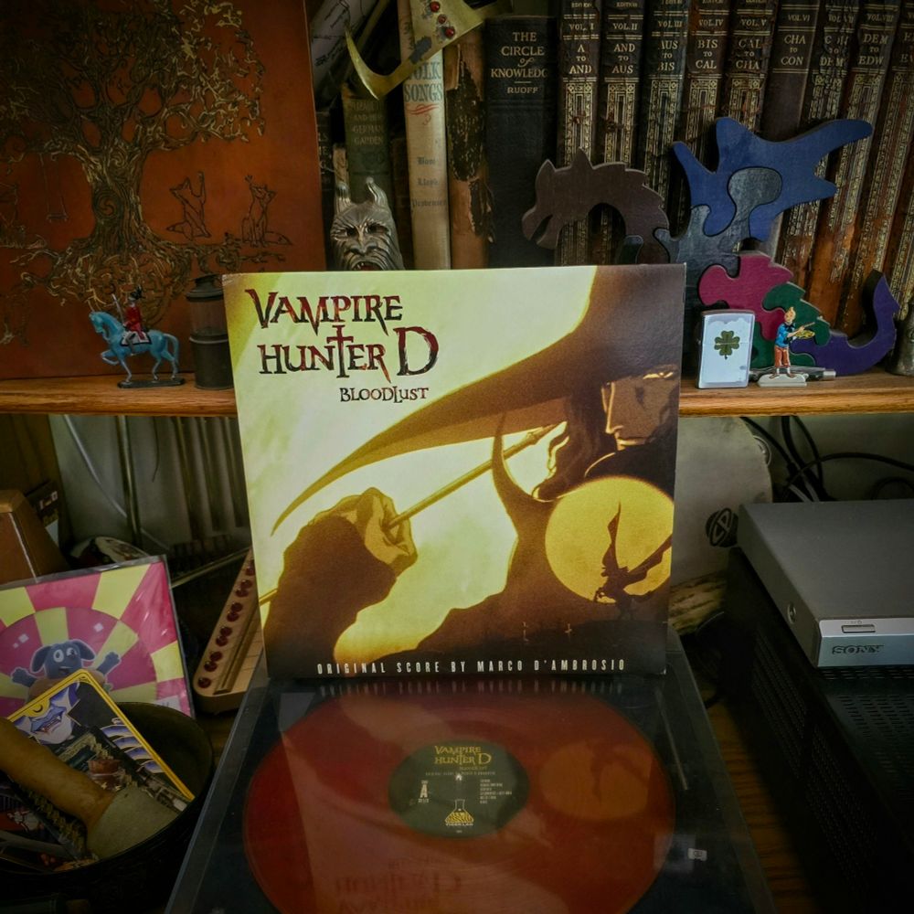 The vinyl album "Vampire Hunter D Bloodlust OST" by Marco D'Ambrosio sitting on a turntable surrounded by the album cover sleeve, antique books, curios, tchotchkes, and ephemera. 