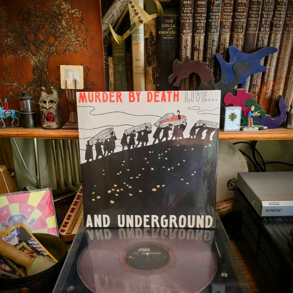 The vinyl album "Live... And Underground" by Murder By Death sitting on a turntable surrounded by the album cover sleeve, antique books, curios, tchotchkes, and ephemera. 