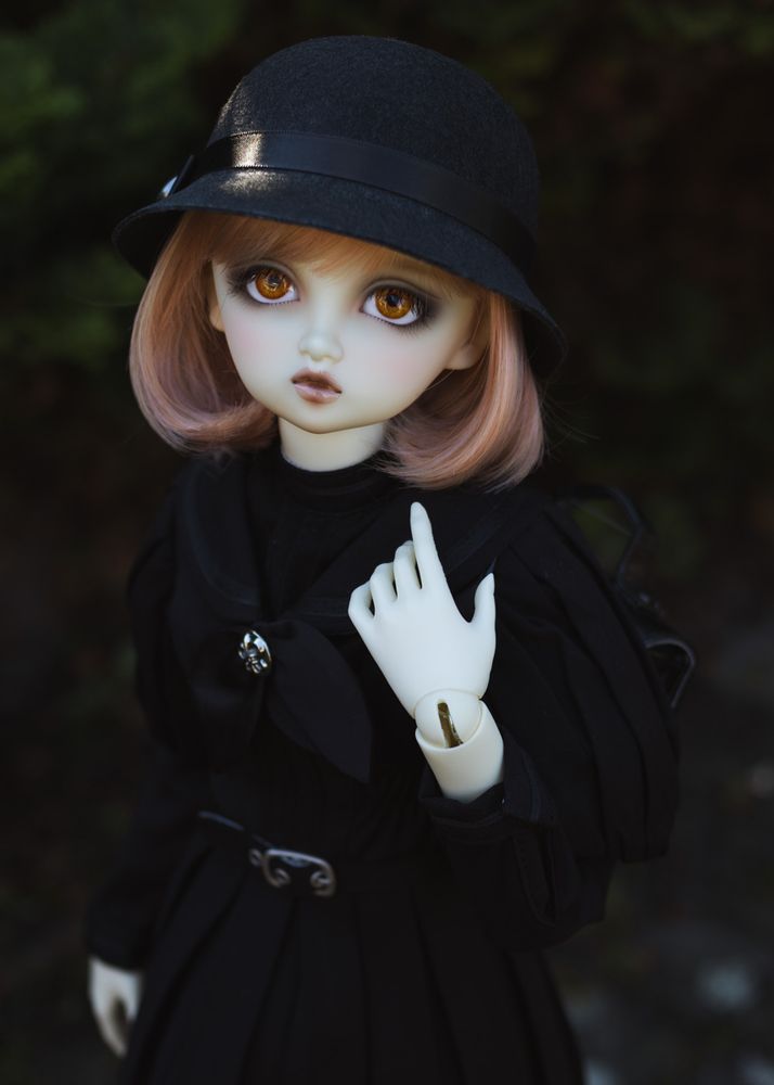 Portrait photo of a ball-jointed resin doll. She is outside in a garden with dark foliage, wearing a black old-fashioned school uniform and a cloche hat. She carries a black school bag on her back. Her resin is pale white, her eyes are orange and her bobbed hair is peach.