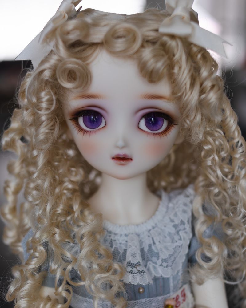 Portrait photo of a ball-jointed doll with very large purple eyes and curly blonde ringlets. She is very pale and has an antique girlish look.