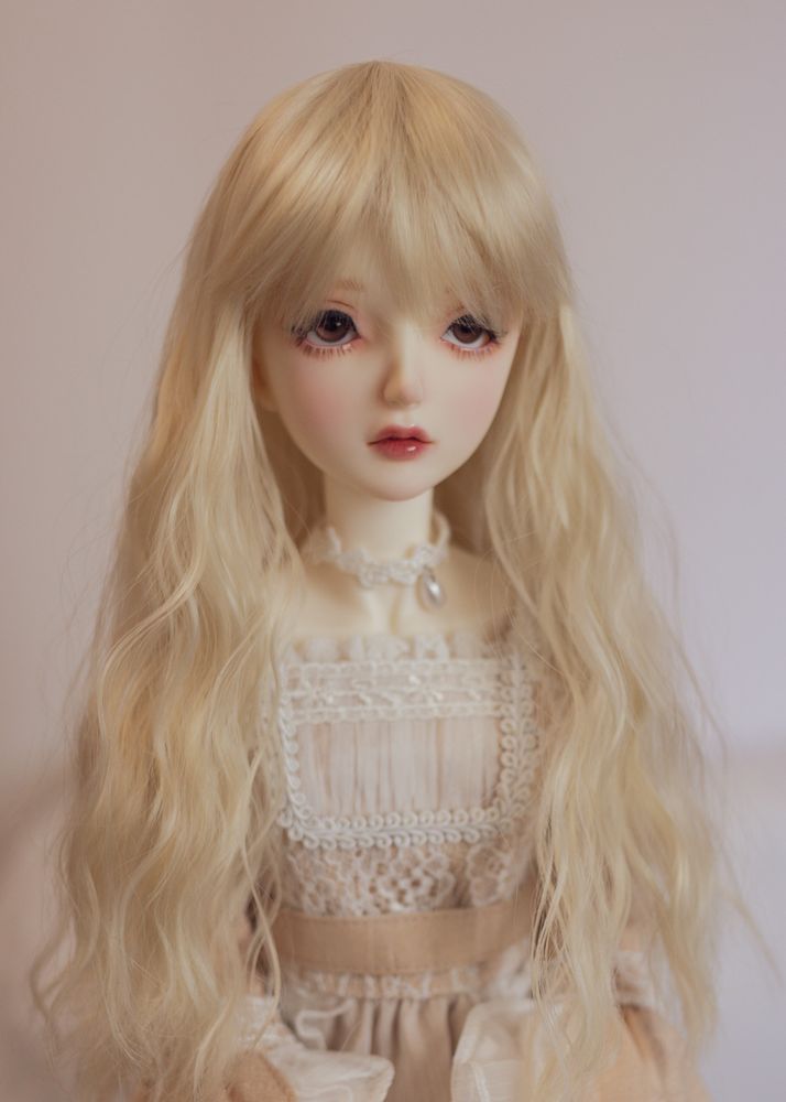 Portrait of a female ball-jointed doll wearing a beige dress decorated with white lace. She has brown eyes and a softly waved pale blonde wig with a long fringe falling over her eyes (or bangs, if you're American!).