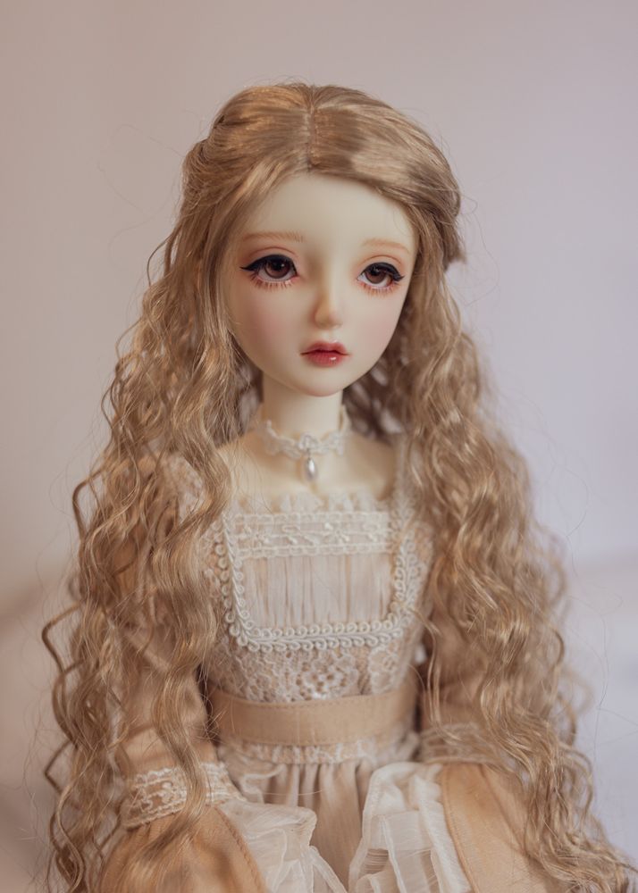 Portrait of a female ball-jointed doll wearing a beige dress decorated with white lace. She has brown eyes and a long, tightly-waved light brown wig, partly pulled back.