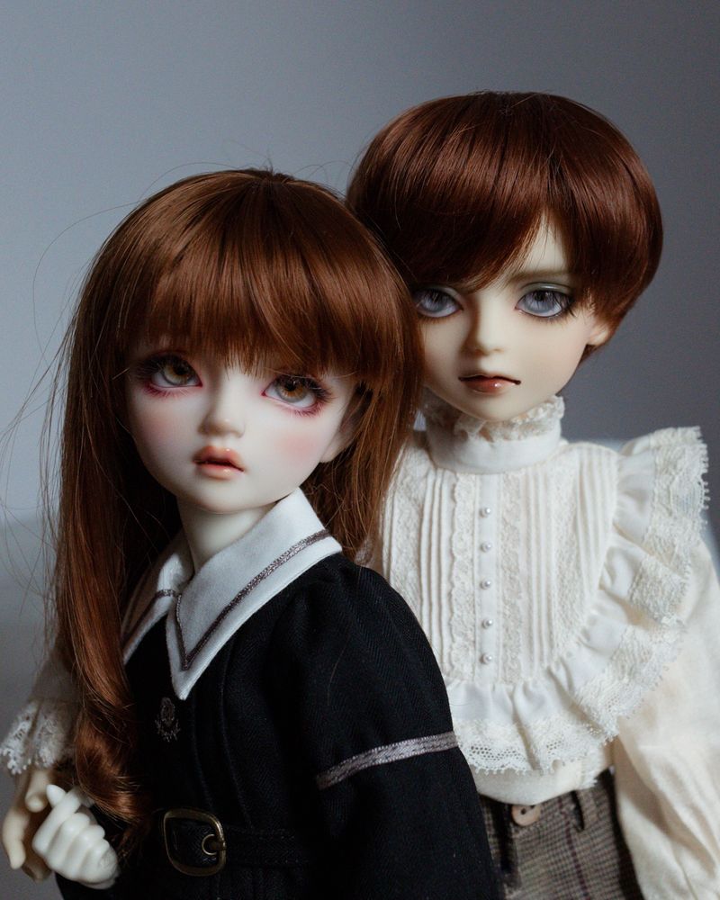 Photo of two resin ball-jointed dolls, a girl (Hazel) and a slightly taller boy (Hewitt), facing slightly away from each other. They both have red-brown hair. The girl wears a black school uniform with a white collar, and the boy wears a very frilly white shirt with lace.