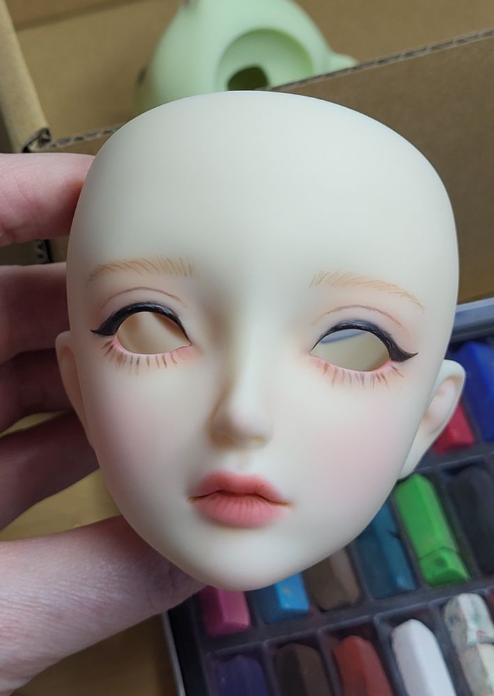 A woman's hand holding the head of a partially painted ball-jointed doll. It has heavy black eyeliner, natural colours in the face, and is a bit messy.
