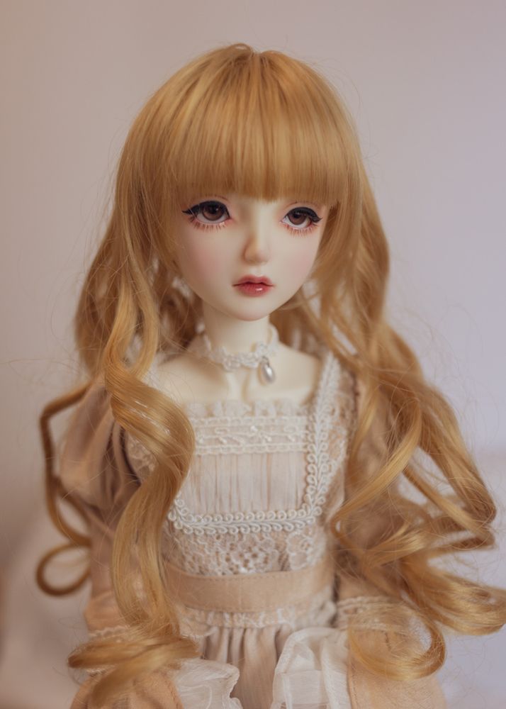 Portrait of a female ball-jointed doll wearing a beige dress decorated with white lace. She has brown eyes and a long, honey blonde wig. It has a long fringe (or bangs, if you're American) and big loose curls.