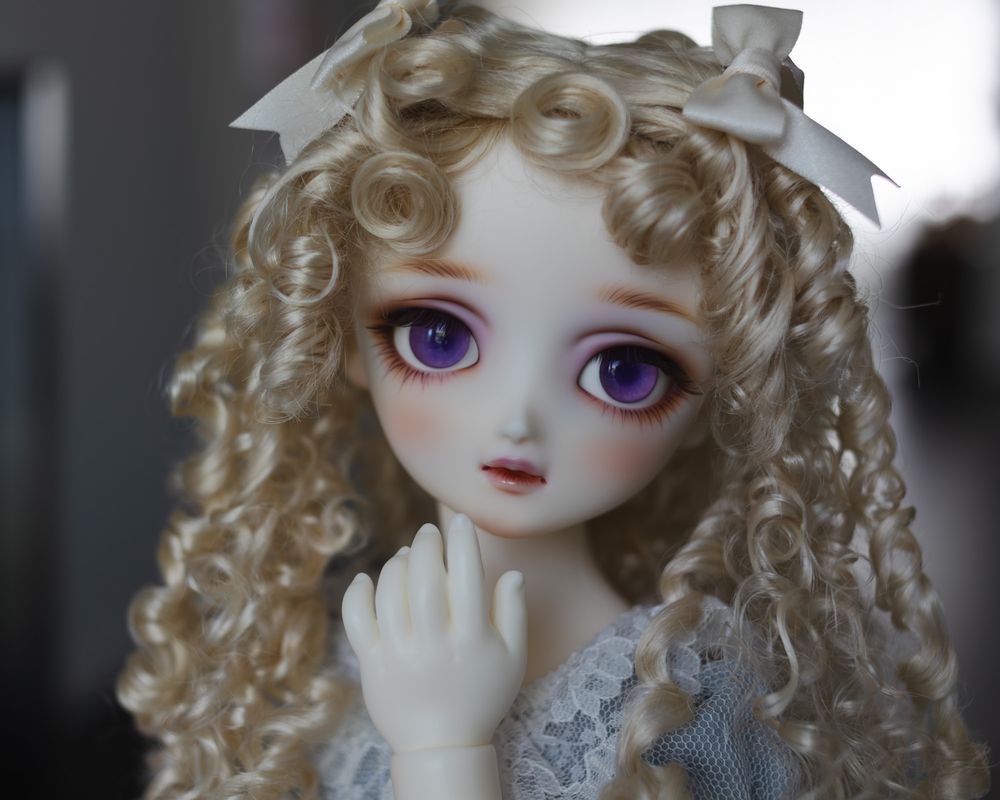 Portrait photo of a ball-jointed doll with very large purple eyes and curly blonde ringlets. She is very pale and has an antique girlish look. Her plump hand is held just below her chin as if she is thinking about something.