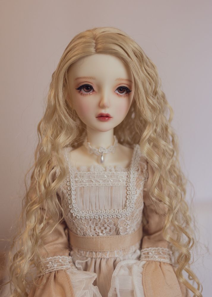 Portrait of a female ball-jointed doll wearing a beige dress decorated with white lace. She has brown eyes and a long, tightly-waved pale blonde wig.