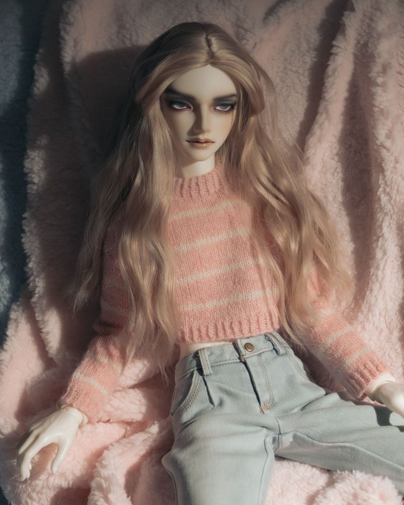 Male ball-jointed doll with pale skin and long ash-pink hair reclines on a pink and blue fur backdrop. He wears a pink and white striped knitted sweater and pale blue jeans, and is looking away from the camera. 