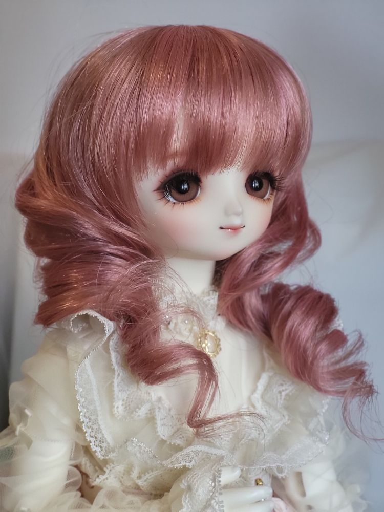 Female ball-jointed doll with a very stylised anime-like face with big brown eyes and a pink wig with a large curl over each shoulder. She is looking away from the camera in a three-quarter portrait view and is wearing an ivory frilly blouse with lace and sheer sleeves.
