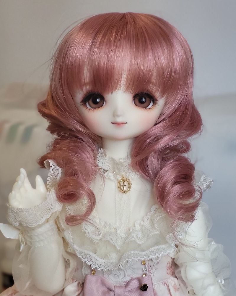Female ball-jointed doll with a very stylised anime-like face with big brown eyes and a pink wig with a large curl over each shoulder. She has a hand raised as if waving at the camera and is wearing an ivory frilly blouse with lace and sheer sleeves.