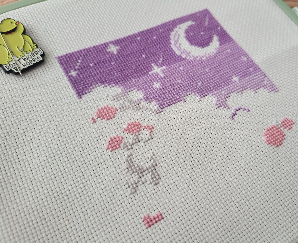 A cross stitch piece in progress on white Aida fabric, with a purple gradient sky with moon and stars, plus the start of some grey and pink clouds. In one corner of the piece is a needle minder. It's got a happy frog on it, with the text "Don't worry be hoppy!"