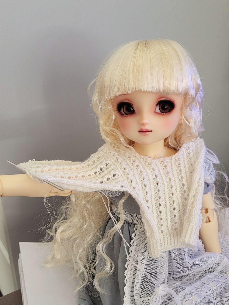 A ball-jointed doll with a cute big-eyed face and a long curly blonde wig is sitting with one arm out. An in-progress white lace knitted jumper is draped over her body and the arm, to check fit.