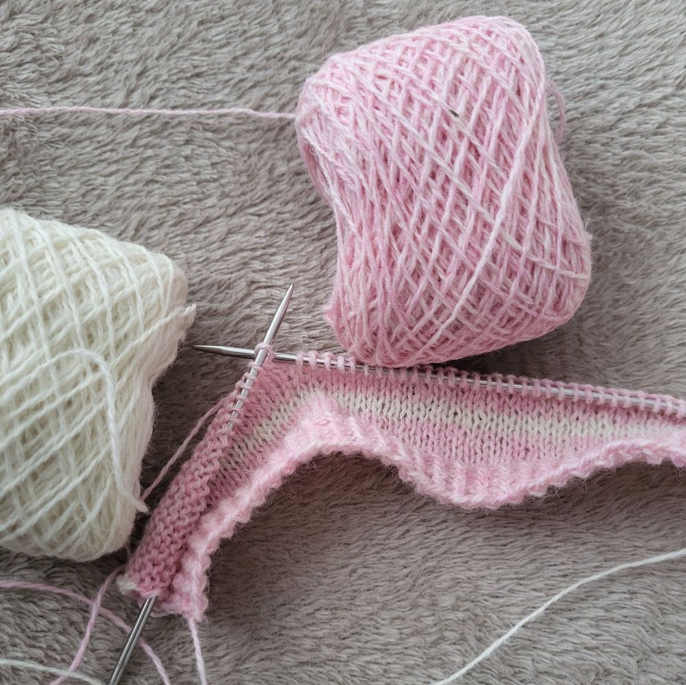A short piece of knitting in progress, still on the knitting needles. The knitted fabric is pale pink with a white stripe. In the background are two balls of wool, one pink and one white. 
