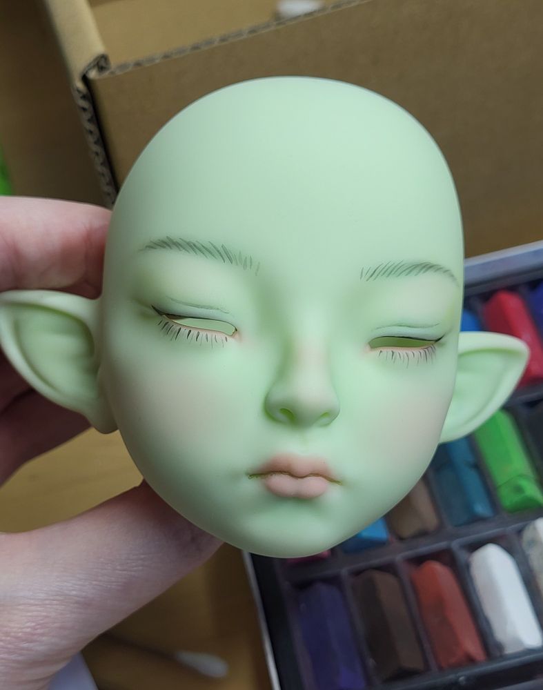 A woman's hand holding the head of a partially painted light green ball-jointed doll. It has pointed ears and half-closed eyes.