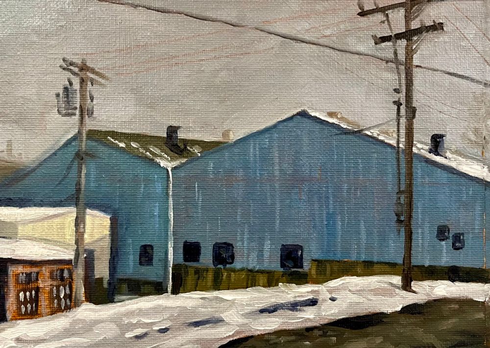 A set of industrial buildings with peaked roofs stand in the snow set against a gray sky and power lines. 