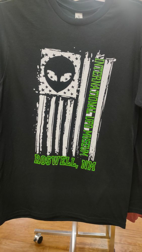 Black t-shirt with screen printed design: black and white version of USA flag in vertical orientation. Superimposed over the field of stars in black is the face of a stereotypical alien (pointed head, large eyes). Green text reading "International UFO Museum" overlays on of the flags stripes. At the bottom of the image is green text that reads, "Roswell, NM" 