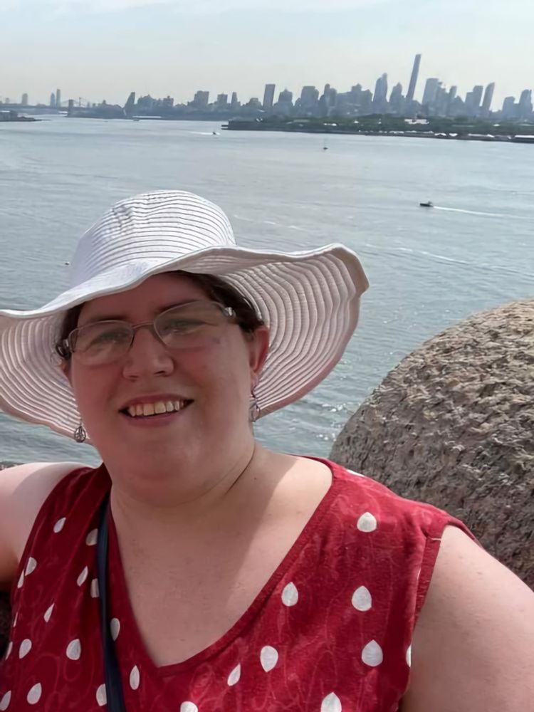 A woman with brown hair and glasses stands in front of a view of NYC and the harbor from the Statue of Liberty base. She is wearing a red shirt with white dots and a white hat.