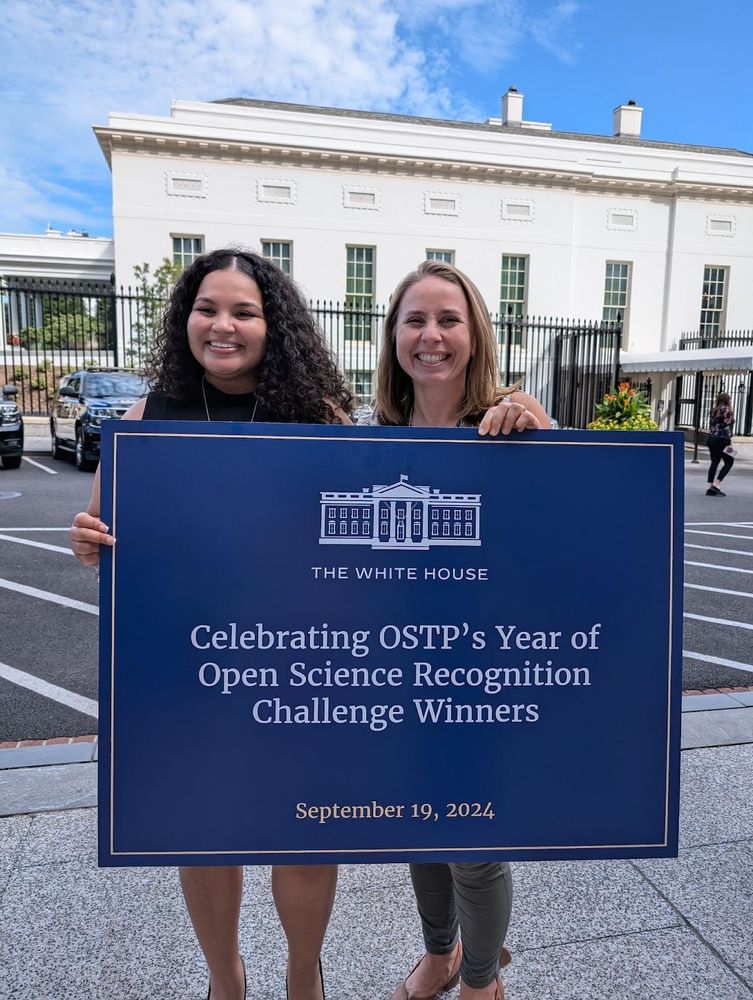 a Black woman and a white woman smiling standing in front of a large single storey building holding a large sign with logo of The White House and text 'Celebrating OSTP's Year of Open Science Recognition Challenge Winners dated September 19, 2024'. Blue sky with white clouds