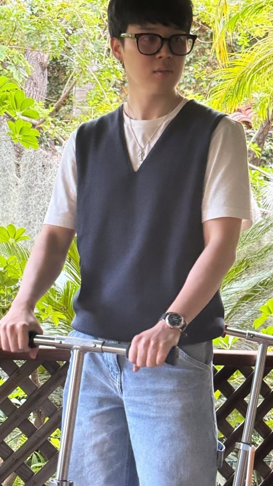 candid jimin photo by taehyung. jimin standing with his hands on the handle bars of a scooter. he's wearing jeans and a white t-shirt with a woollen vest and black framed glasses looking cute. 